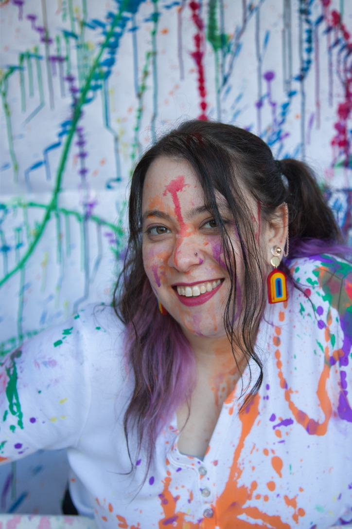 Head and shoulders of a young white woman with long brown hair. Her white shirt and face are splattered with multicoloured paint, and she is wearing a rainbow earring.